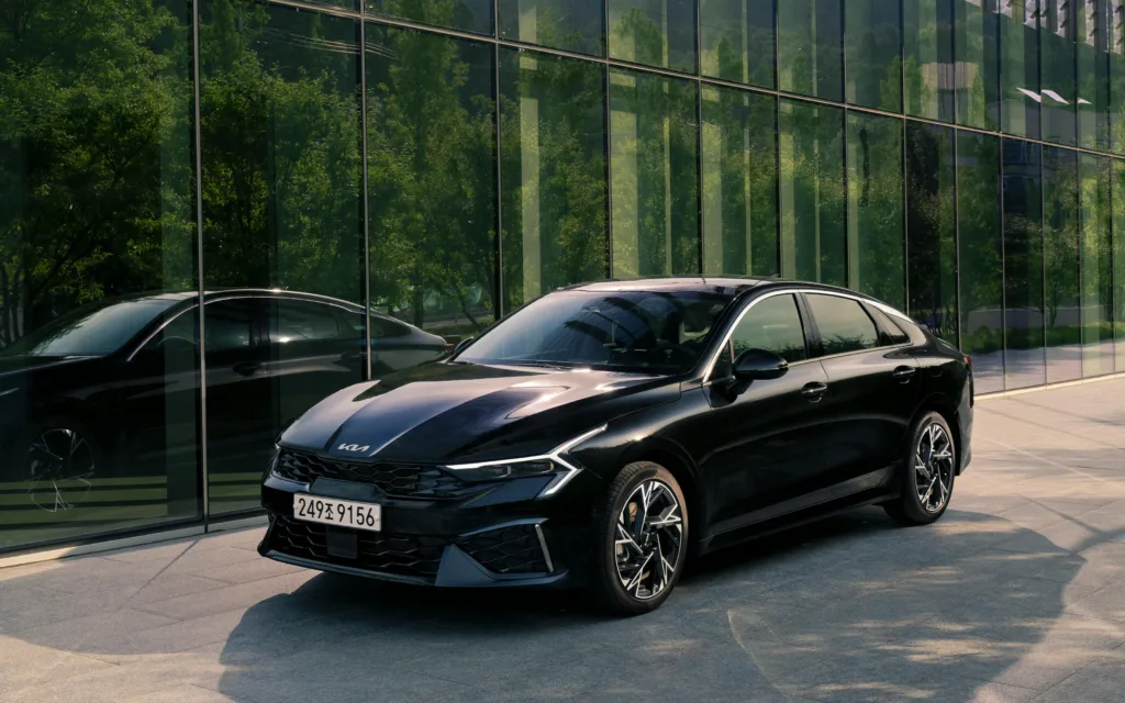 The left front view of the Kia K5 stopped in front of a green glass building.